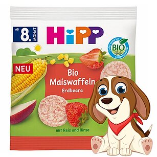 Je zobrazeno balení bio dětské výživy s kukuřičným klasem, jahodou a plátkem jablka, které doprovází kreslený pes.