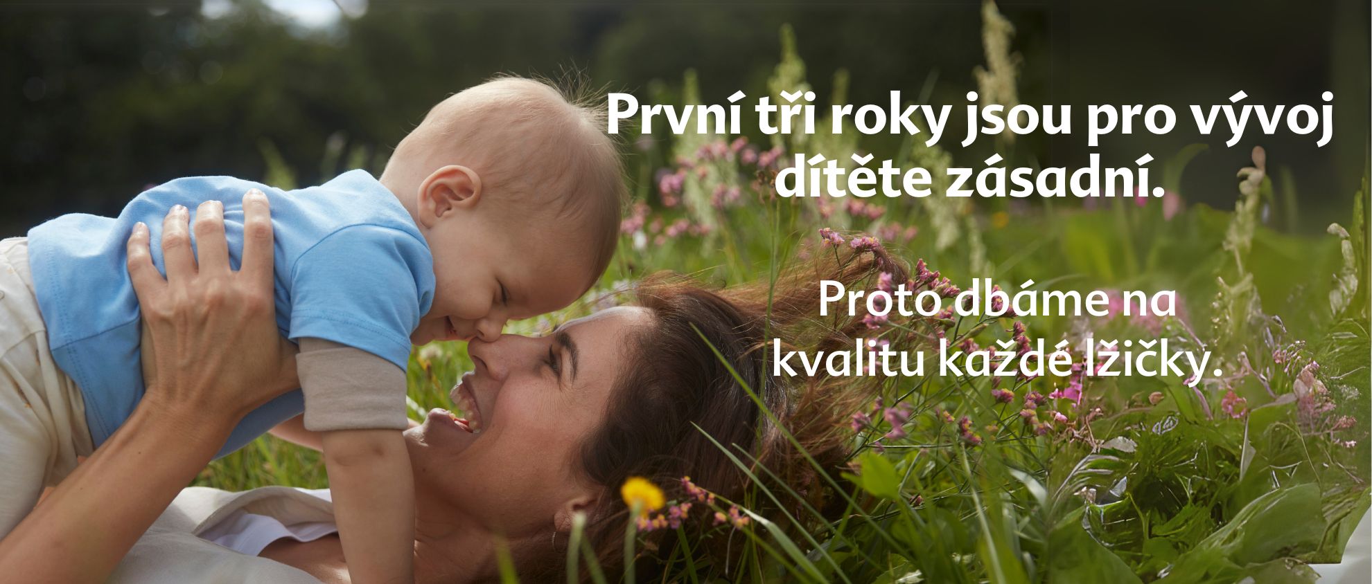 Žena a dítě leží na trávě obklopené květinami a žena se dívá na dítě.