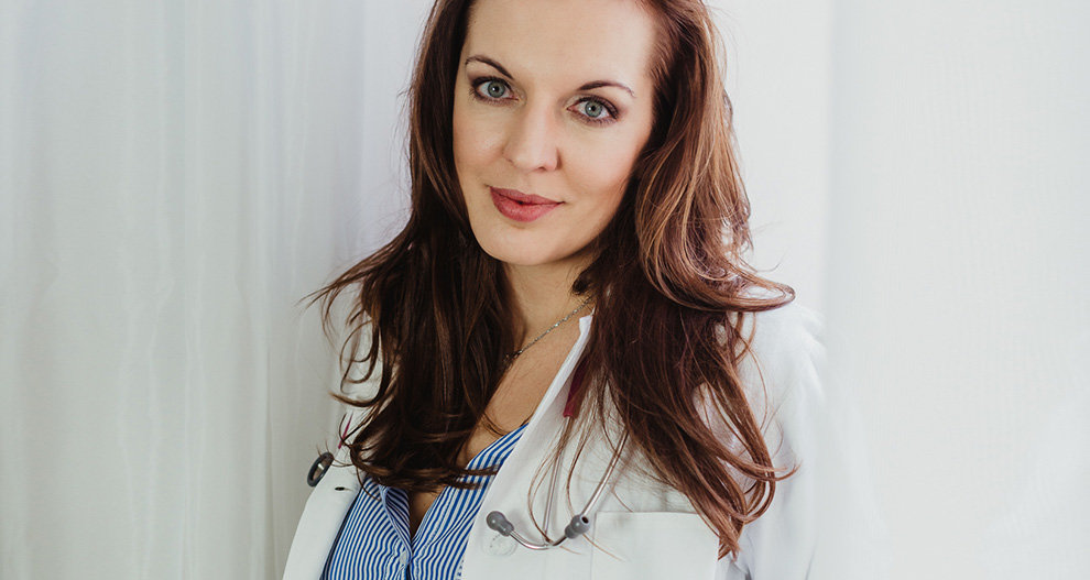 Žena s dlouhými hnědými vlasy, v bílém laboratorním plášti a modré košili, stojí na pozadí bílého závěsu.