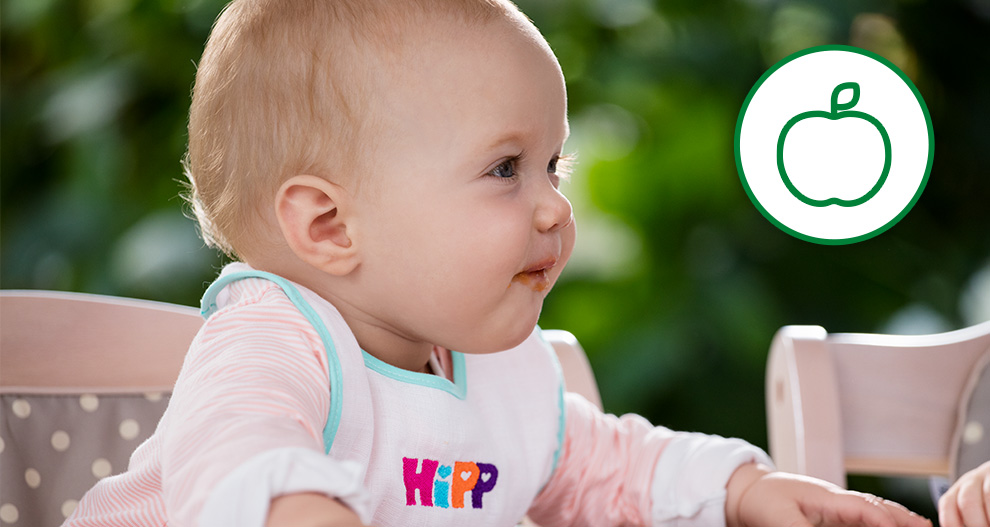 Dítě v židličce s bryndáčkem s nápisem "HIPP" se dívá do strany a na obrázku je zelený kruh s ikonou bílého jablka.