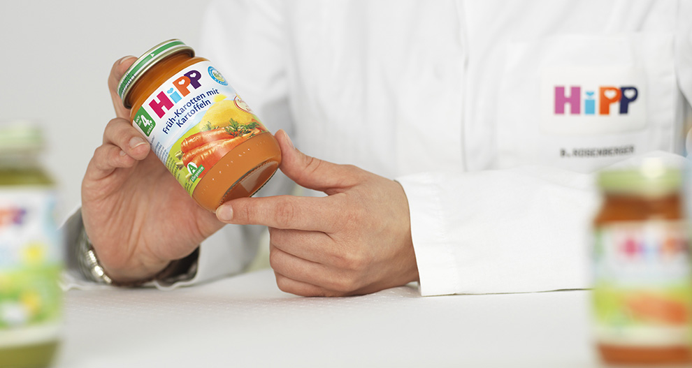 Osoba v bílém laboratorním plášti drží sklenici s dětskou výživou s etiketou "HiPP" a "Karotten-Sauce mit Apfel" (Mrkvová omáčka s jablkem).