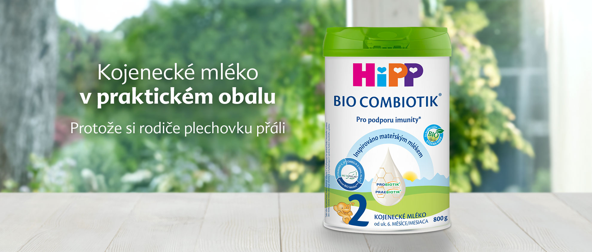 Bílá válcovitá nádoba se zelenými a modrými štítky s ilustrací dítěte a textem "Hipp Bio Combiotik" je umístěna na dřevěném povrchu s rozmazaným pozadím zeleně.