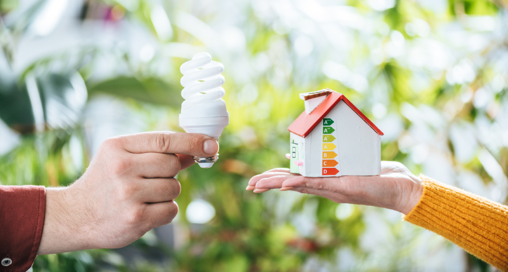 Ruka drží spirálovou kompaktní zářivku, zatímco druhá ruka představuje model domu s hodnocením energetické účinnosti.