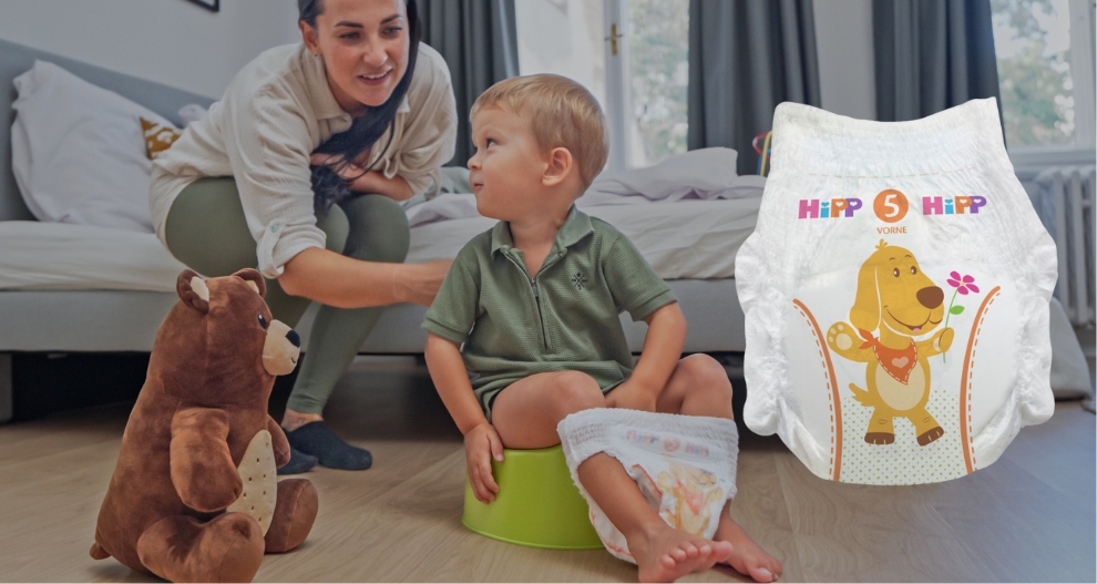 Žena pomáhá malému chlapci se zeleným nočníkem, zatímco v popředí je medvídek a plenka se vzorem psa.
