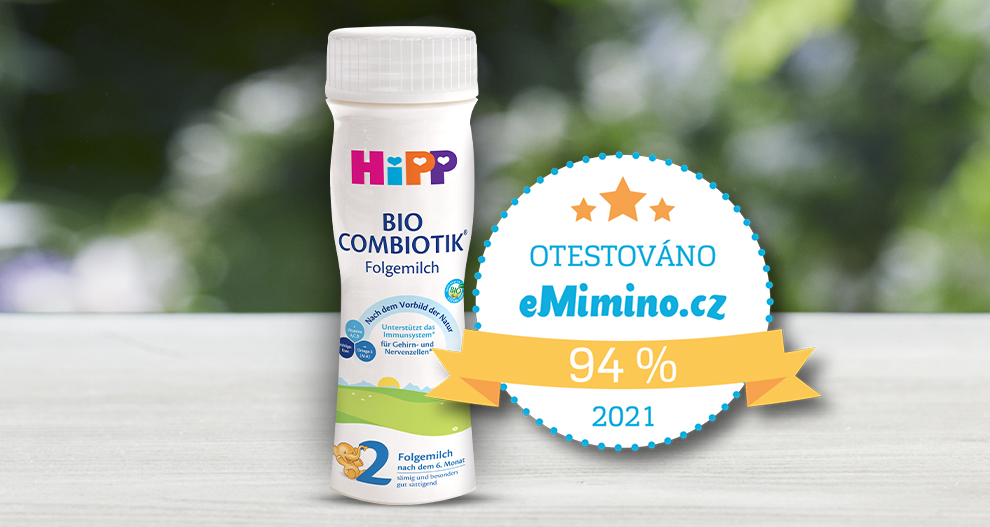 Bílá láhev s nápisem "HiPP Bio Combiotik Folgemilch" s motivem zelené krajiny a ilustrací dítěte je umístěna na dřevěné ploše vedle odznaku s textem "OTESTOVANO eMino.cz 94% 2021".