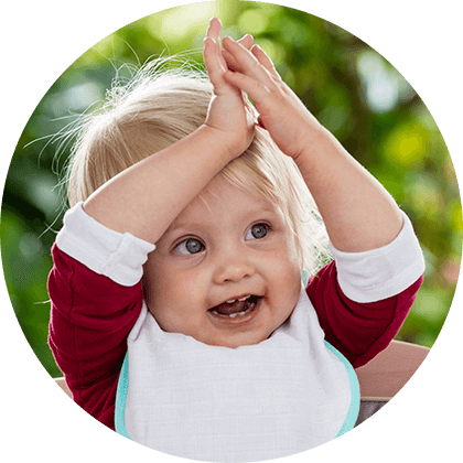 A child with blonde hair, wearing a red shirt and white bib, is raising their hands above their head.