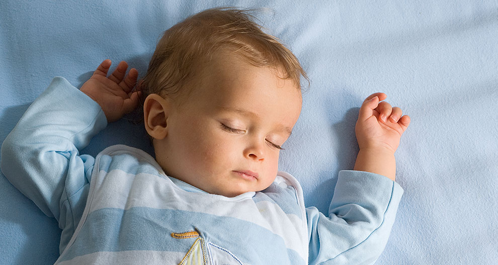 A baby lies on a blue surface, wearing a light blue and white striped shirt with a sailboat design.