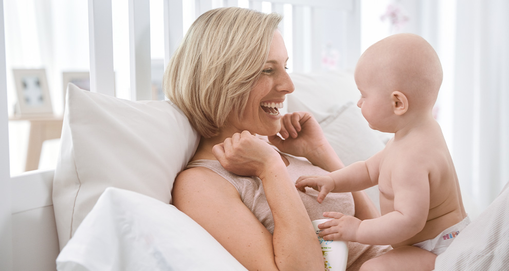 A woman and a baby are lying on a bed, with the woman holding a bottle and the baby reaching out to her.