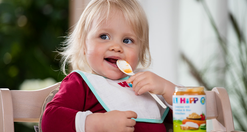 A toddler with blonde hair is seated in a high chair, wearing a red shirt and bib, eating from a jar of Hipp baby food.
