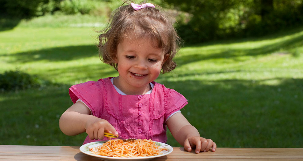 A child in a pink shirt is eating spaghetti from a white plate outdoors.