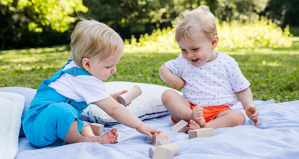 Two toddlers sit on a blanket outdoors, playing with wooden blocks.