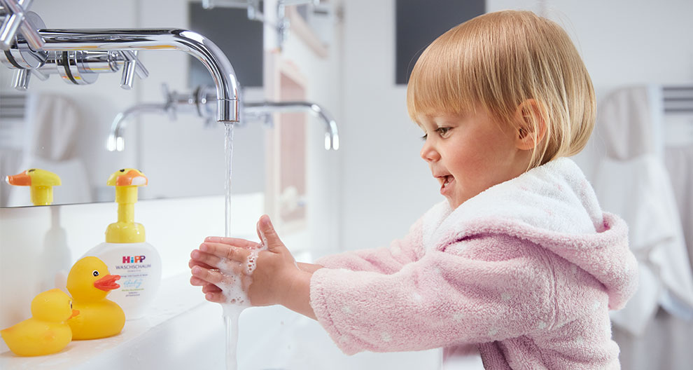 A child in a pink robe washes hands under a faucet with a yellow rubber duck toy beside a bottle of soap.