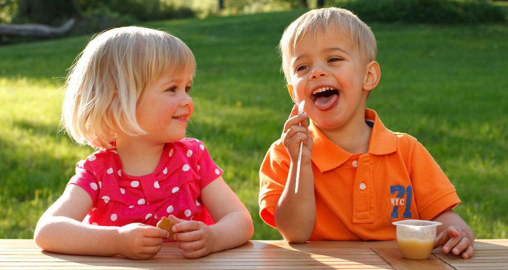 Two children, a girl in a pink dress and a boy in an orange shirt, sit at a table outdoors, with the girl holding a cookie and the boy eating from a cup.