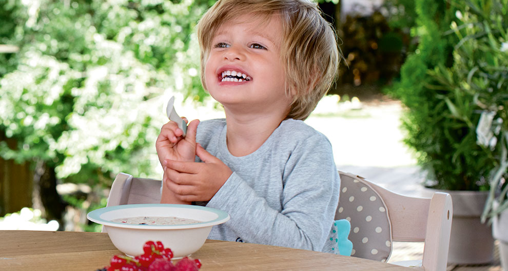 A child is seated at a table with a bowl of food and a spoon, looking up with a smile.